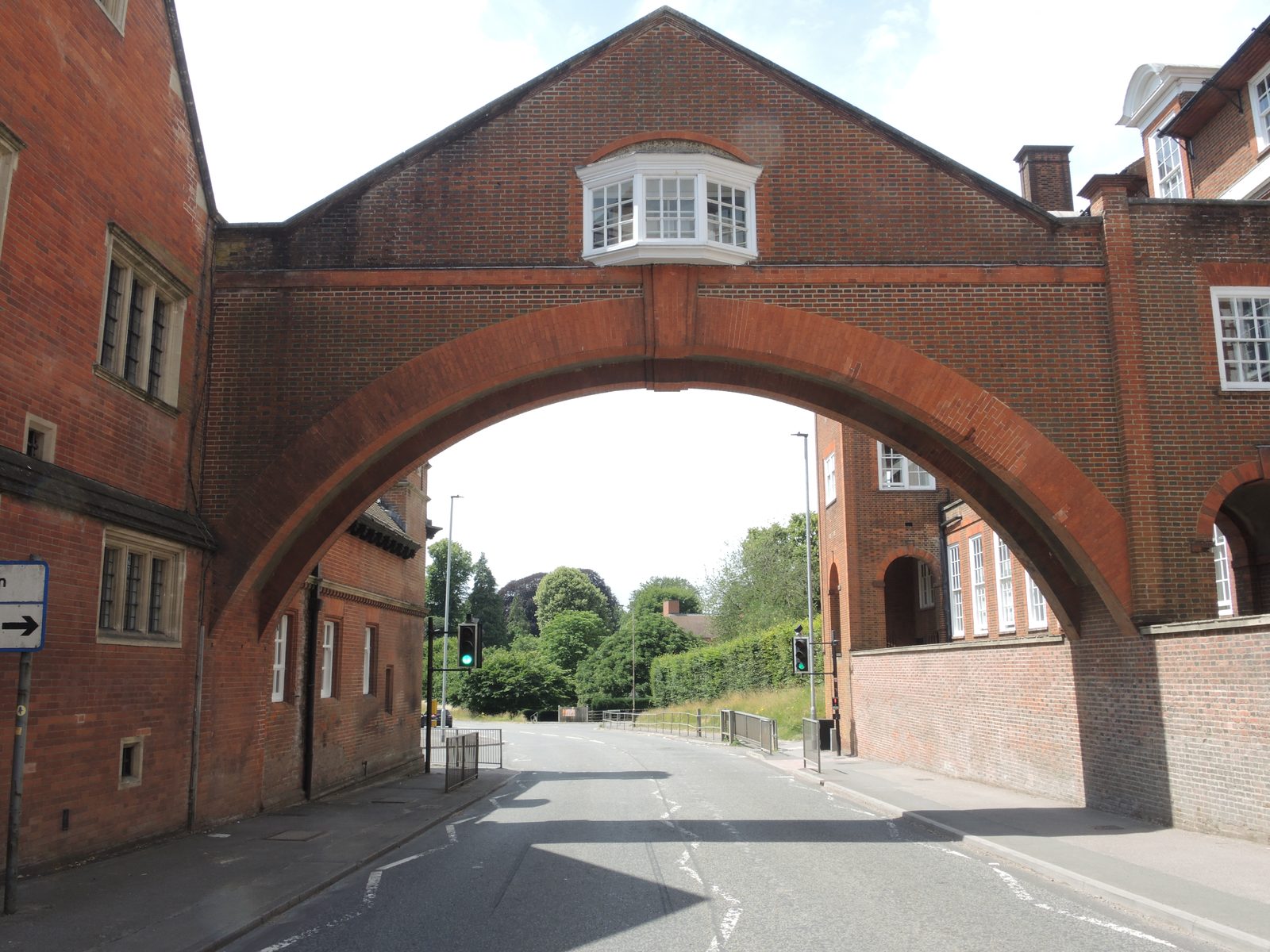 The Marlborough College footbridge over the A4 Bath Road: an arched twentieth-century pedestrian bridge in red brick and white stone dressings, spanning the main road between the college grounds and the Marlborough Mound side, with mature trees framing the view from the road.